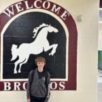 A school hallway wall features a large maroon, black, and white Broncos welcome mural beside interior windows with blinds, with a student standing in front.