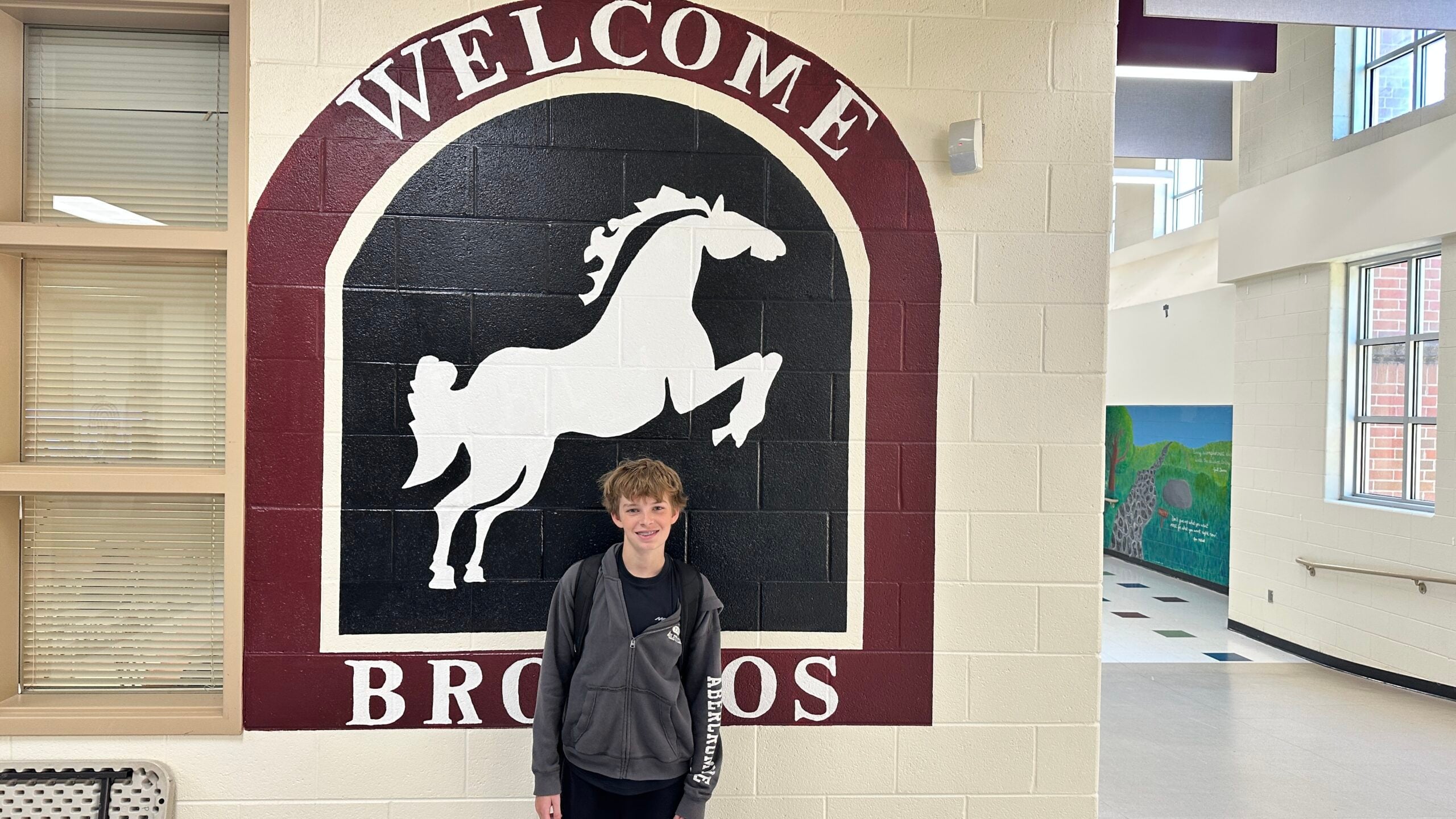 A school hallway wall features a large maroon, black, and white Broncos welcome mural beside interior windows with blinds, with a student standing in front.